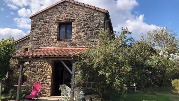 A stone building with a tiled roof, a small window, and a covered porch with a chair. - Gite Daim, Gite en Pleine Nature (Avène)