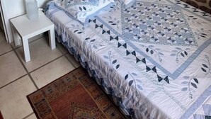 A bedroom with a stone wall, a white bed with patterned bedding, a small bedside table, and a nightstand. - Gite Daim, Gite en Pleine Nature (Avène)