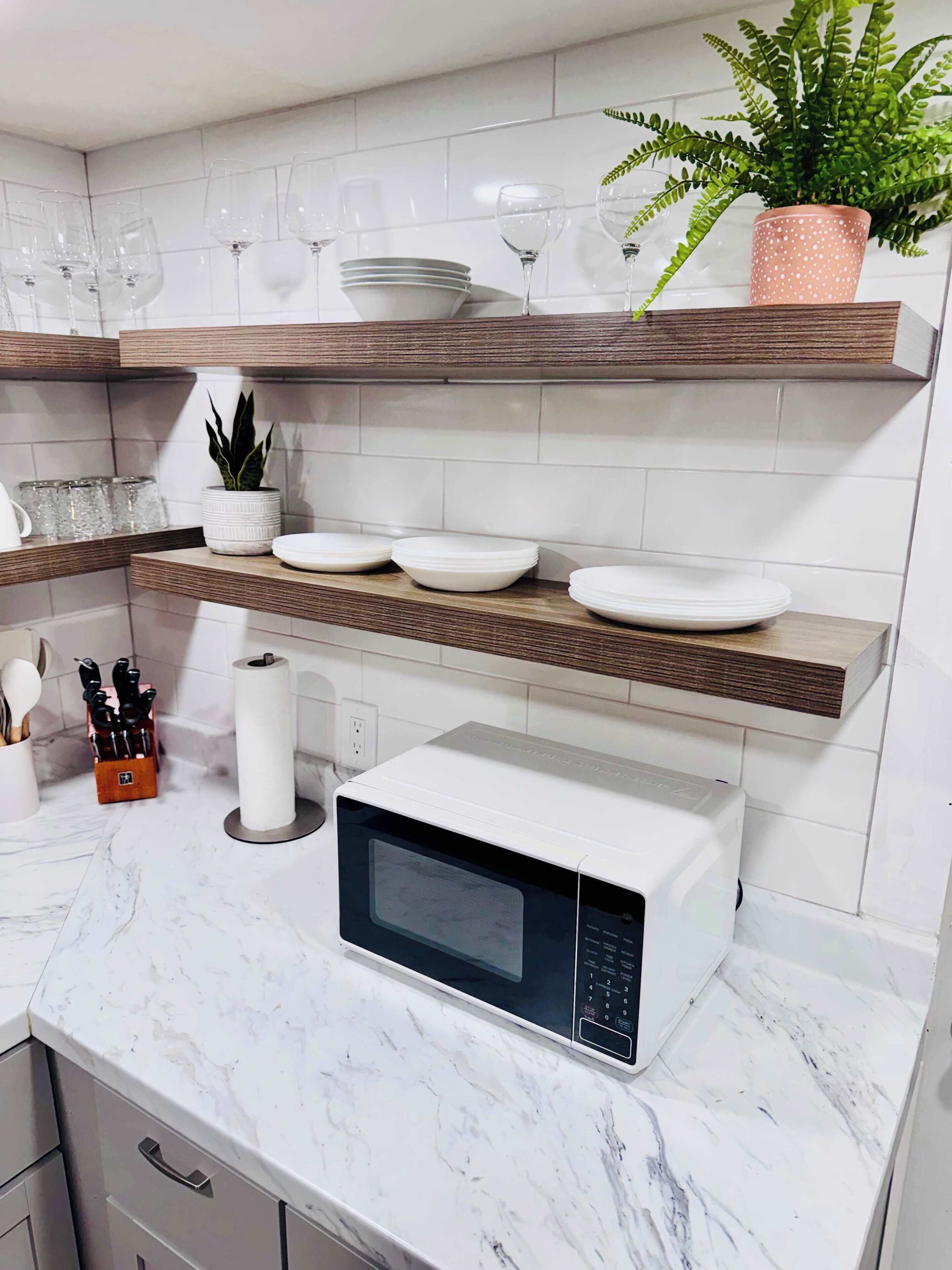 A kitchen with white marble countertops, a microwave, a potted plant, and neatly arranged dishes and glasses on wooden shelves.