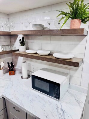 Una cocina con encimeras de mármol blanco, microondas, una planta en maceta y vajilla y cristalería ordenadas en estanterías de madera.
