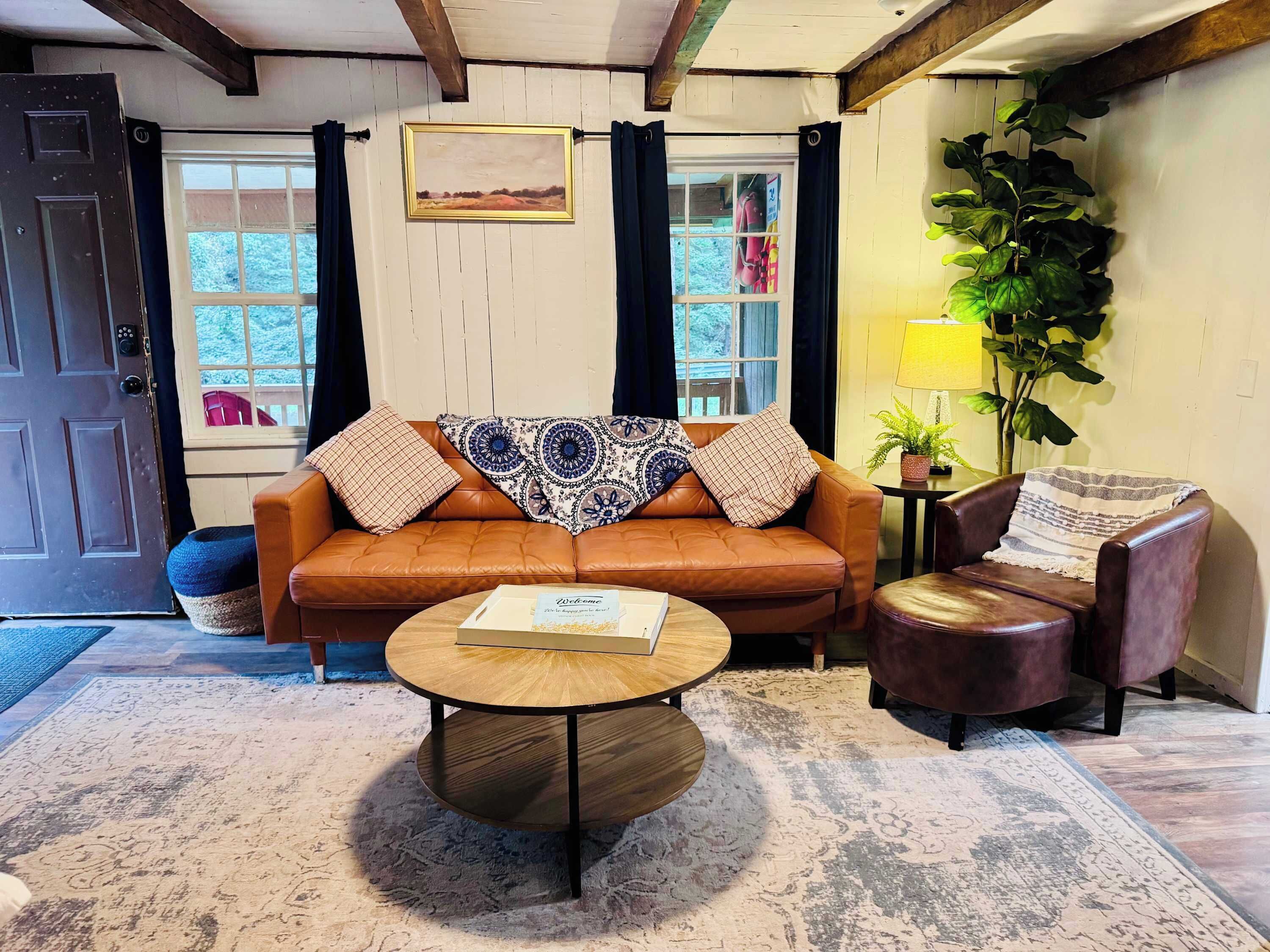 A living room with a brown leather sofa, a round wooden coffee table, and a brown leather armchair. There is a window with curtains, a framed picture on the wall, and a potted plant.