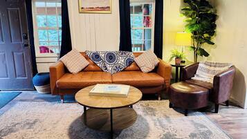 A living room with a brown leather sofa, a round wooden coffee table, and a brown leather armchair. There is a window with curtains, a framed picture on the wall, and a potted plant.