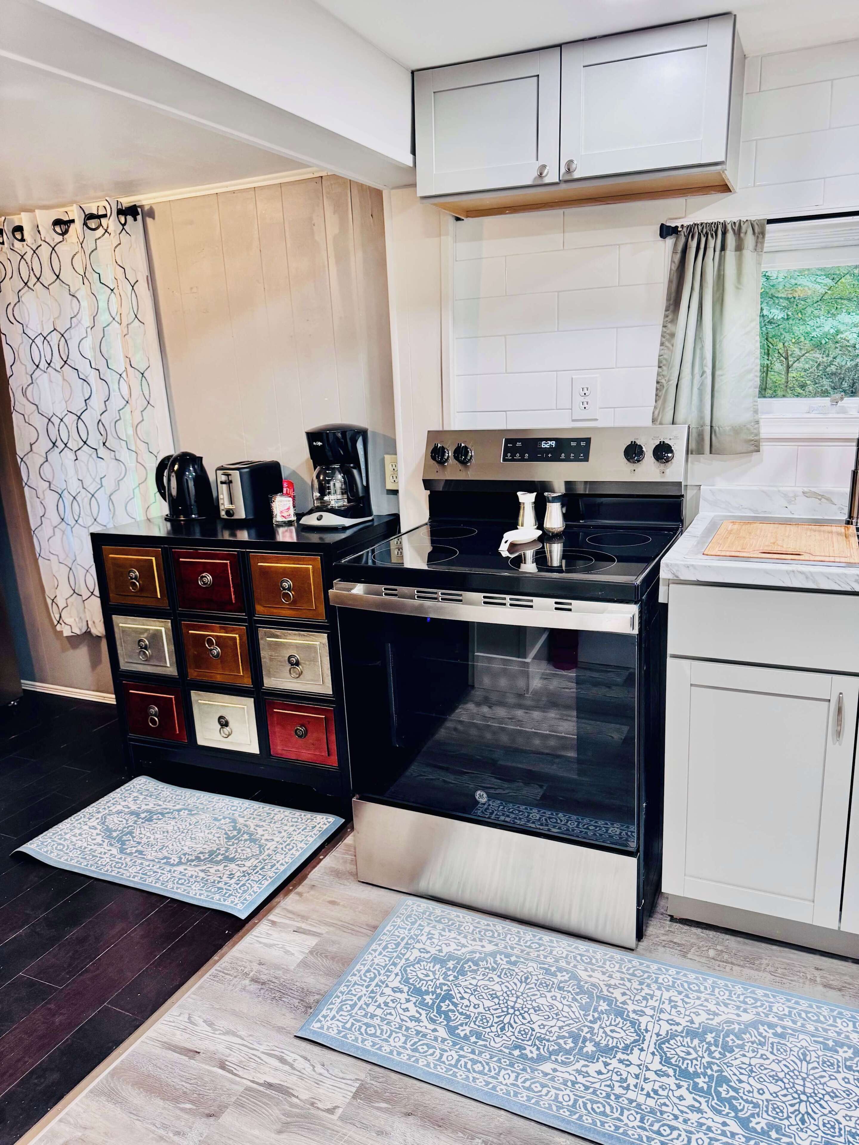 A compact kitchen with a stove, oven, and a coffee maker. There are cabinets and a window with curtains.