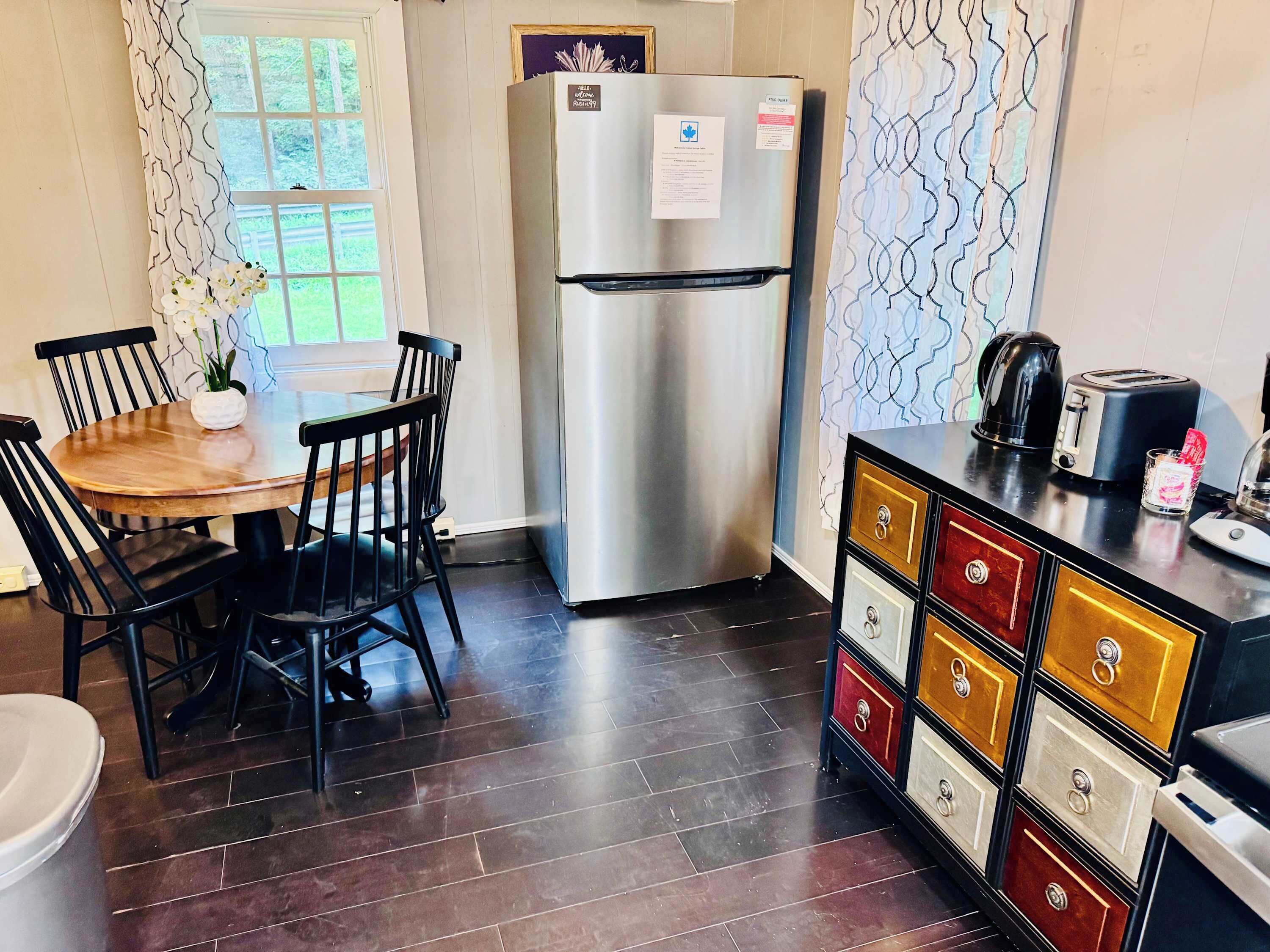A compact kitchen with a stainless steel refrigerator, a wooden dining table with chairs, and a cabinet with multiple drawers.