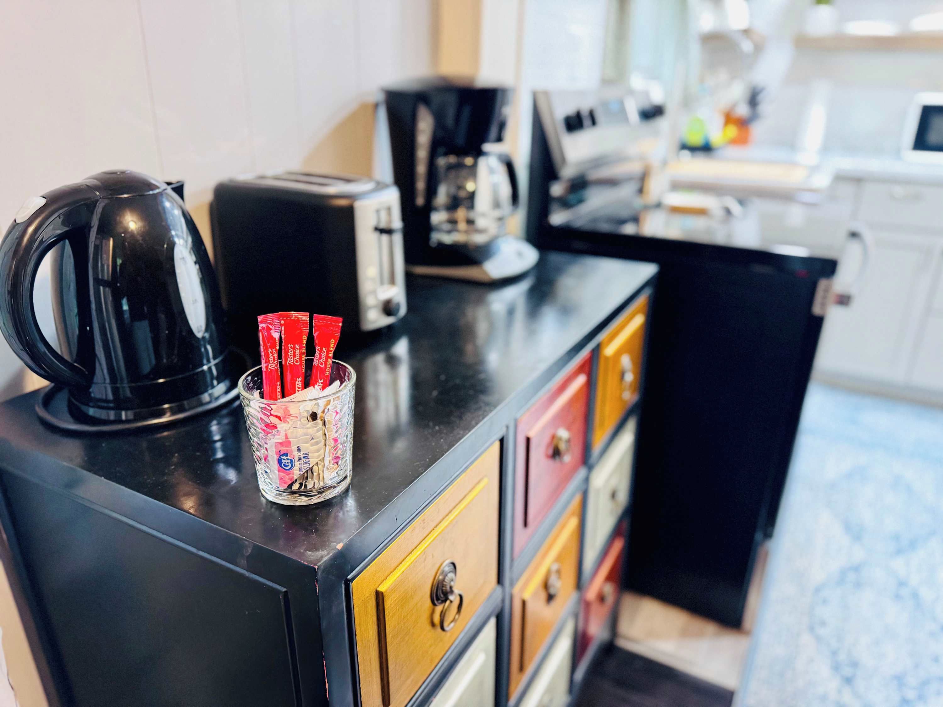 Een kitchenette met een koffiezetapparaat, toaster en een glazen bak met rode zakjes op een zwart aanrecht.