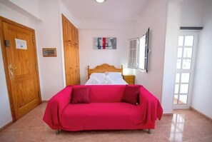 A bedroom with a bed, a wooden wardrobe, a red sofa, a painting on the wall, and a window. - Apartamento Estudio Mirador de los Poetas (Puerto de las Nieves)