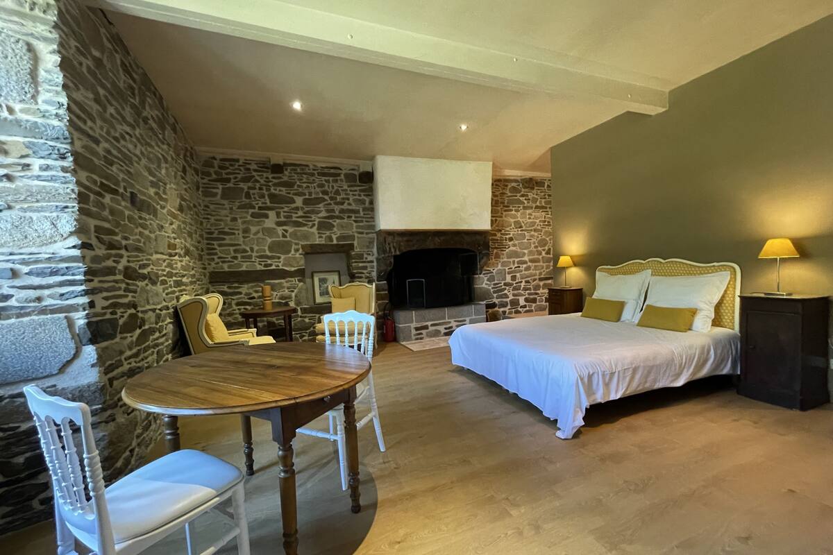 Een kamer met een stenen muur, een open haard, een bed met wit beddengoed en een eettafel met stoelen.