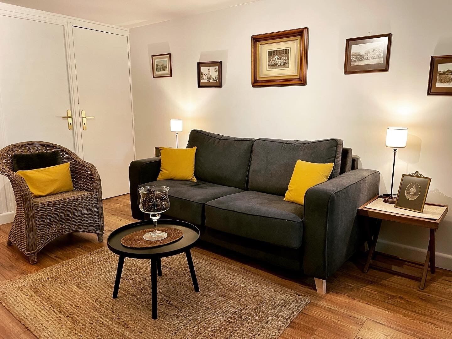 A living room with a dark grey sofa, two yellow cushions, a wicker chair, a small round table with a glass top, and a floor lamp.