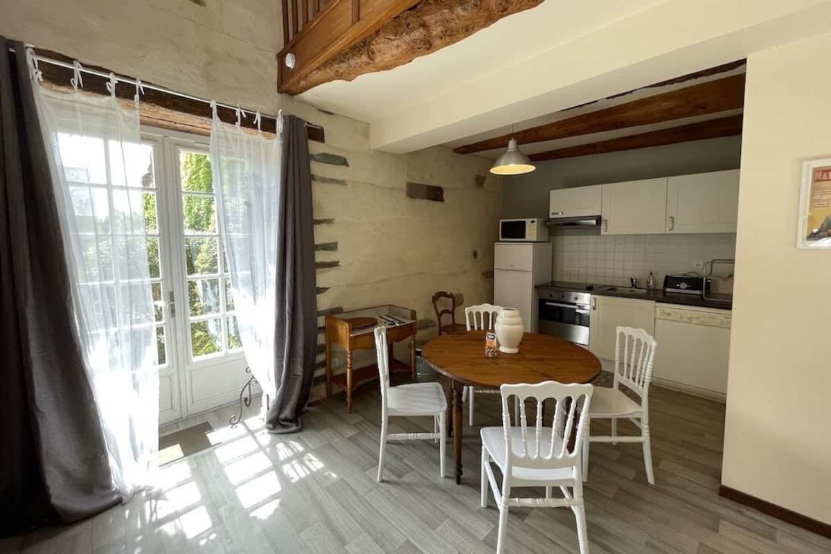 Een eetgedeelte met een houten tafel en witte stoelen, een keuken met witte kasten en apparatuur, en een stenen muur.