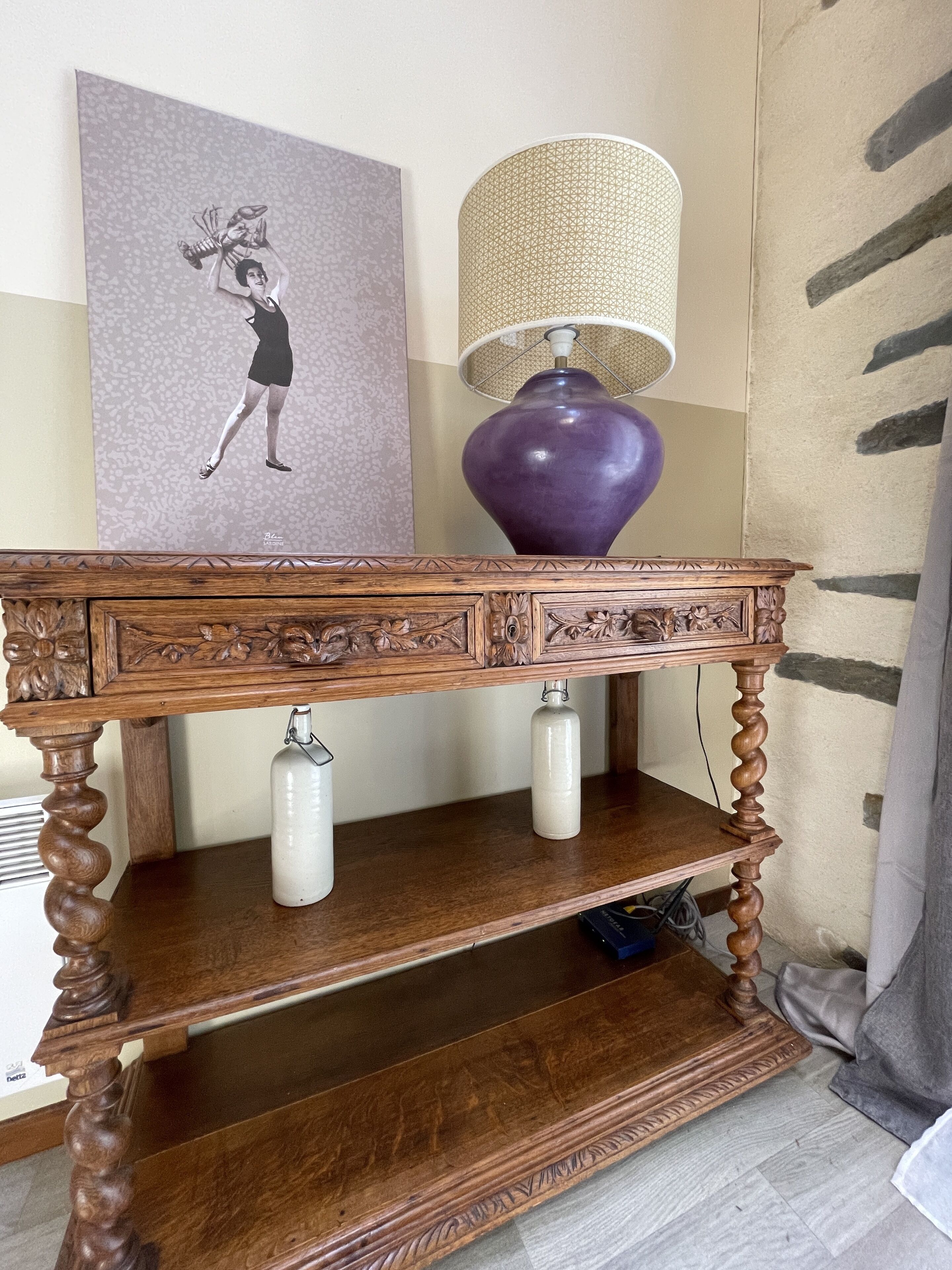 A wooden console table with intricate carvings, a purple lamp, and a framed artwork on the wall.