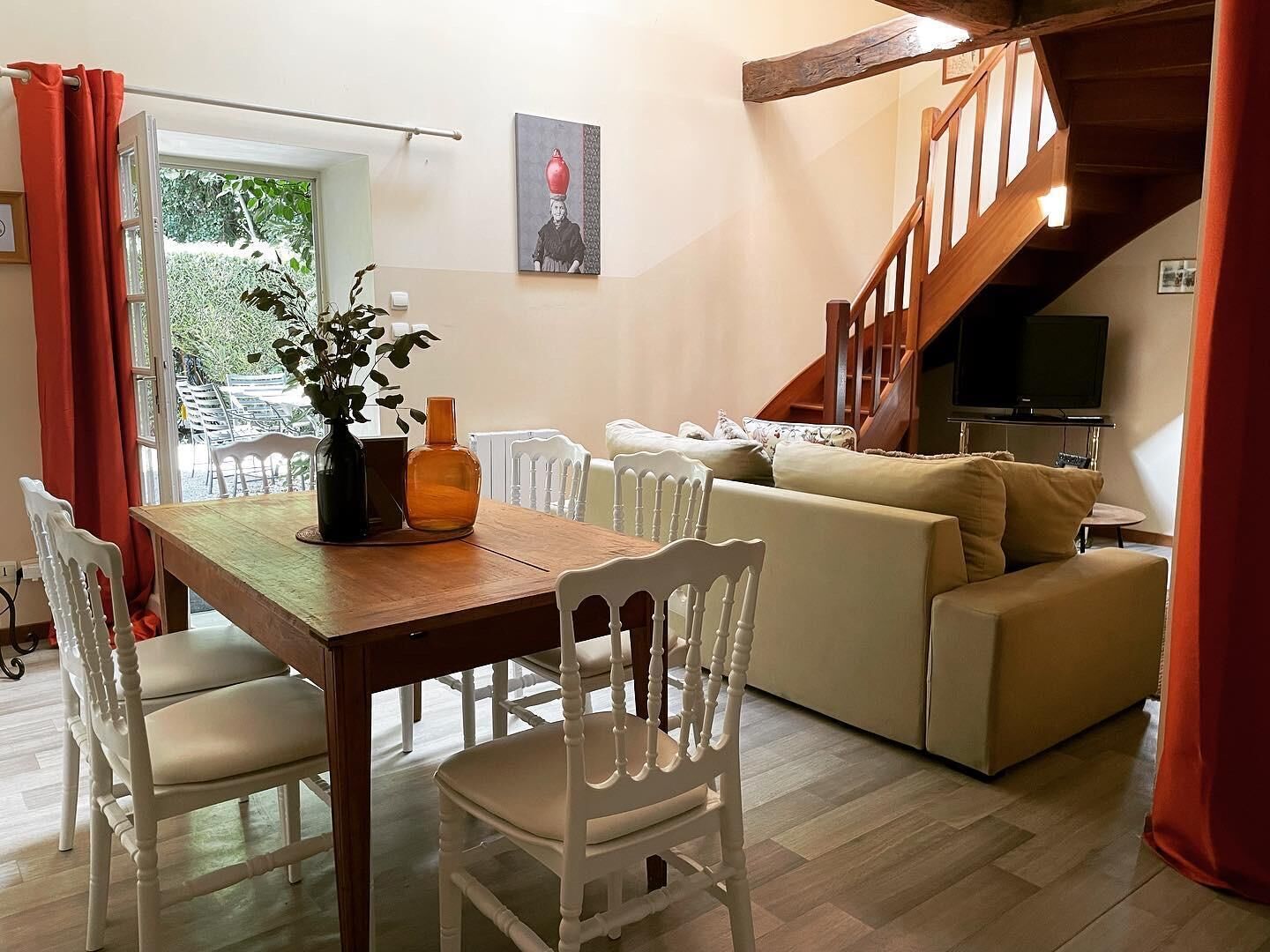 Un coin repas avec une table en bois et des chaises blanches, un canapé beige et un escalier avec un téléviseur.