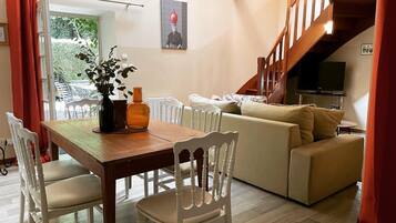 Un coin repas avec une table en bois et des chaises blanches, un canapé beige et un escalier avec un téléviseur.