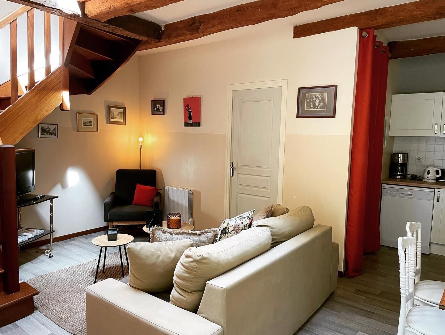A cozy living room with a sofa, a chair, a small table, and a TV. There's a staircase with wooden beams and a red curtain in the kitchen area.