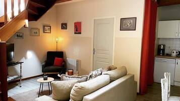 A cozy living room with a sofa, a chair, a small table, and a TV. There's a staircase with wooden beams and a red curtain in the kitchen area.