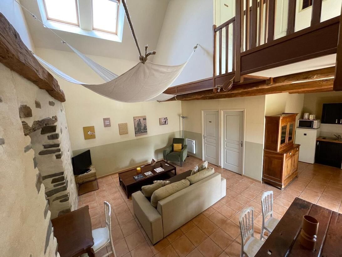 Amplia sala de estar con una hamaca, un sofá, una mesa de centro y una mesa de comedor. Se observa una escalera que conduce a un nivel superior, y un área de cocina al fondo.