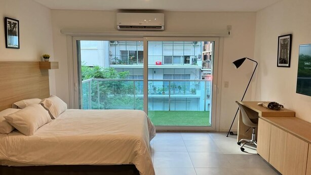 Un dormitorio moderno con cama, escritorio y vista a un balcón con vegetación.