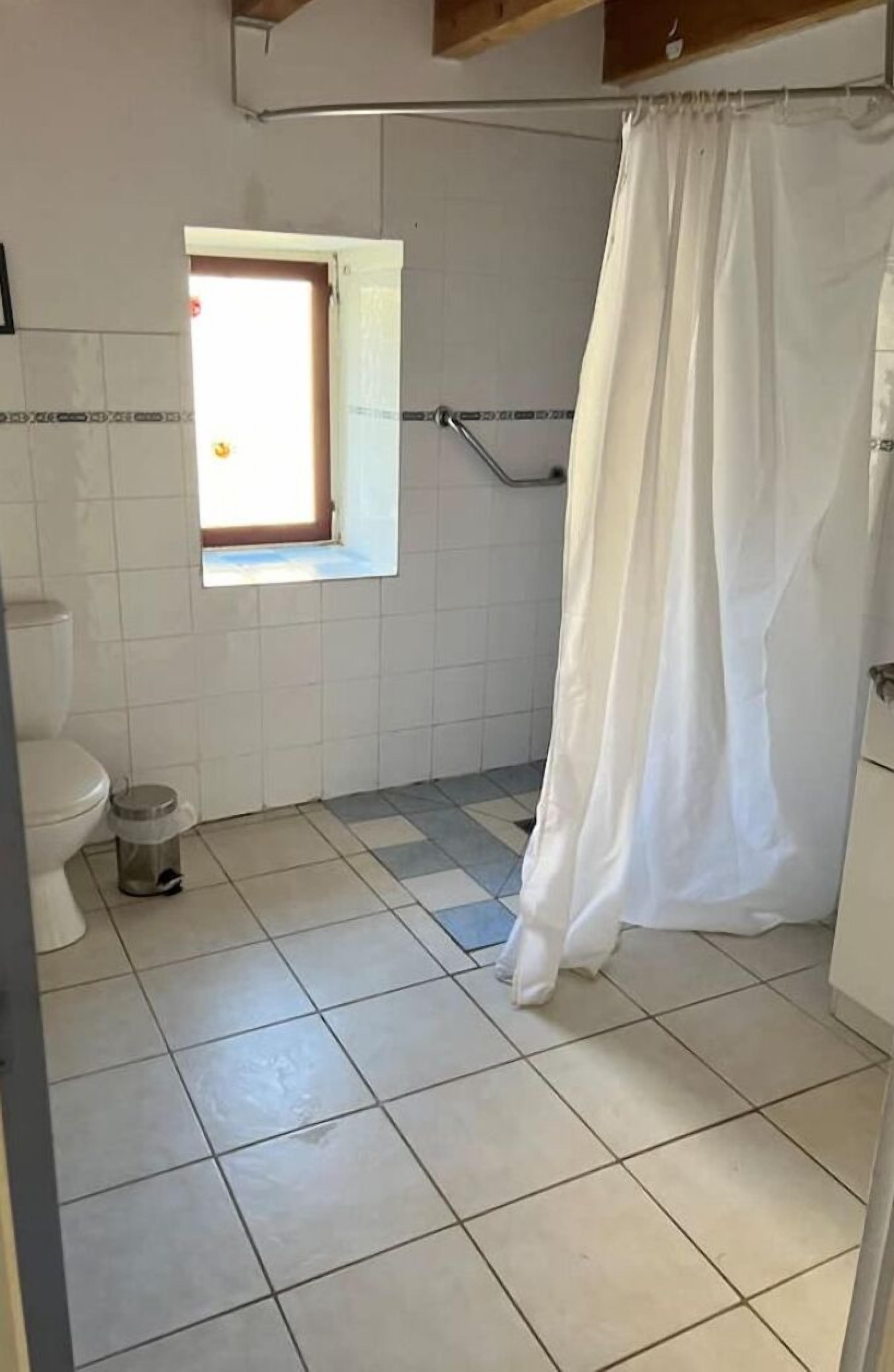 A bathroom with a toilet, a window, a shower curtain, and a trash bin.