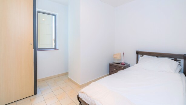 Un dormitorio con cama, mesita de noche, lámpara y ventana.