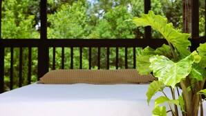 Un lit avec un drap blanc et un oreiller beige, une grande plante verte au premier plan, et une terrasse en bois avec vue sur les arbres à l’arrière-plan.