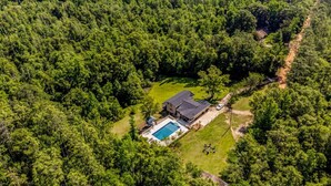 Vue aérienne d’une maison entourée d’une forêt dense, avec une piscine et une terrasse dallée.