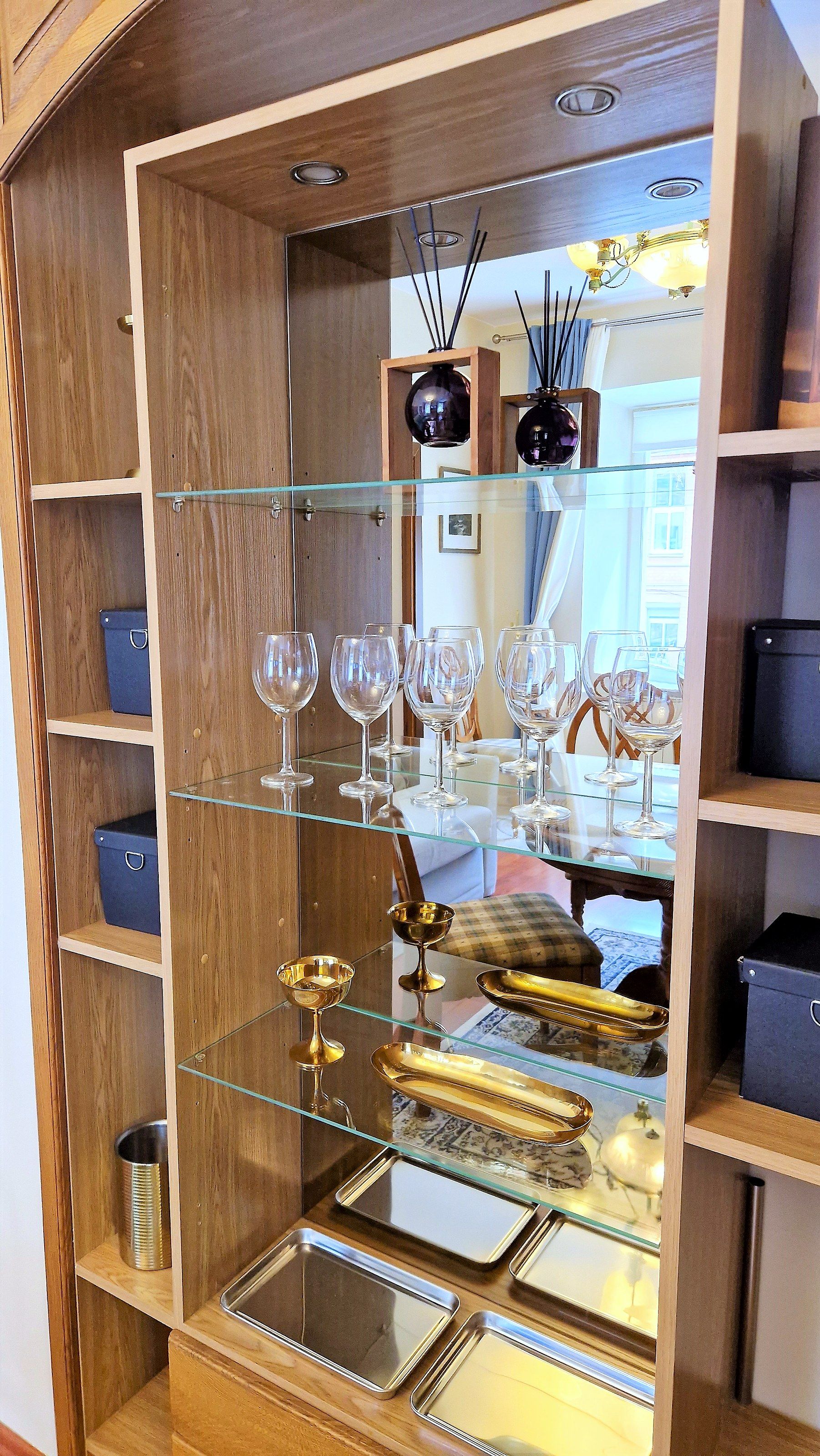 A wooden cabinet with glass shelves holding wine glasses, a gold goblet, and trays.