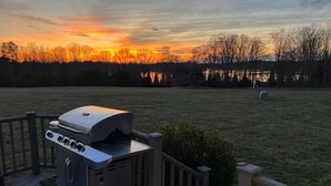 A barbecue grill on a wooden deck with a view of a sunset over a grassy field and trees. - The Hanger House on Lake Norman Airpark (Mooresville)