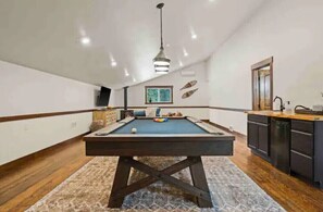 A pool table in a room with wooden flooring, a ceiling light, and a kitchen area with cabinets.