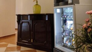 Un área de recepción con piso a cuadros, mostrador de madera oscura, máquina expendedora, vitrina de vidrio y un cuadro de un castillo en la pared.