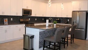 A modern kitchen with white cabinets, a central island with bar stools, a stainless steel refrigerator, and a black tiled backsplash. - Champions Family Home - Near Disney (Davenport)