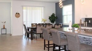 A modern kitchen with a central island, dining area with chairs, and a large window with blinds. - Champions Family Home - Near Disney (Davenport)