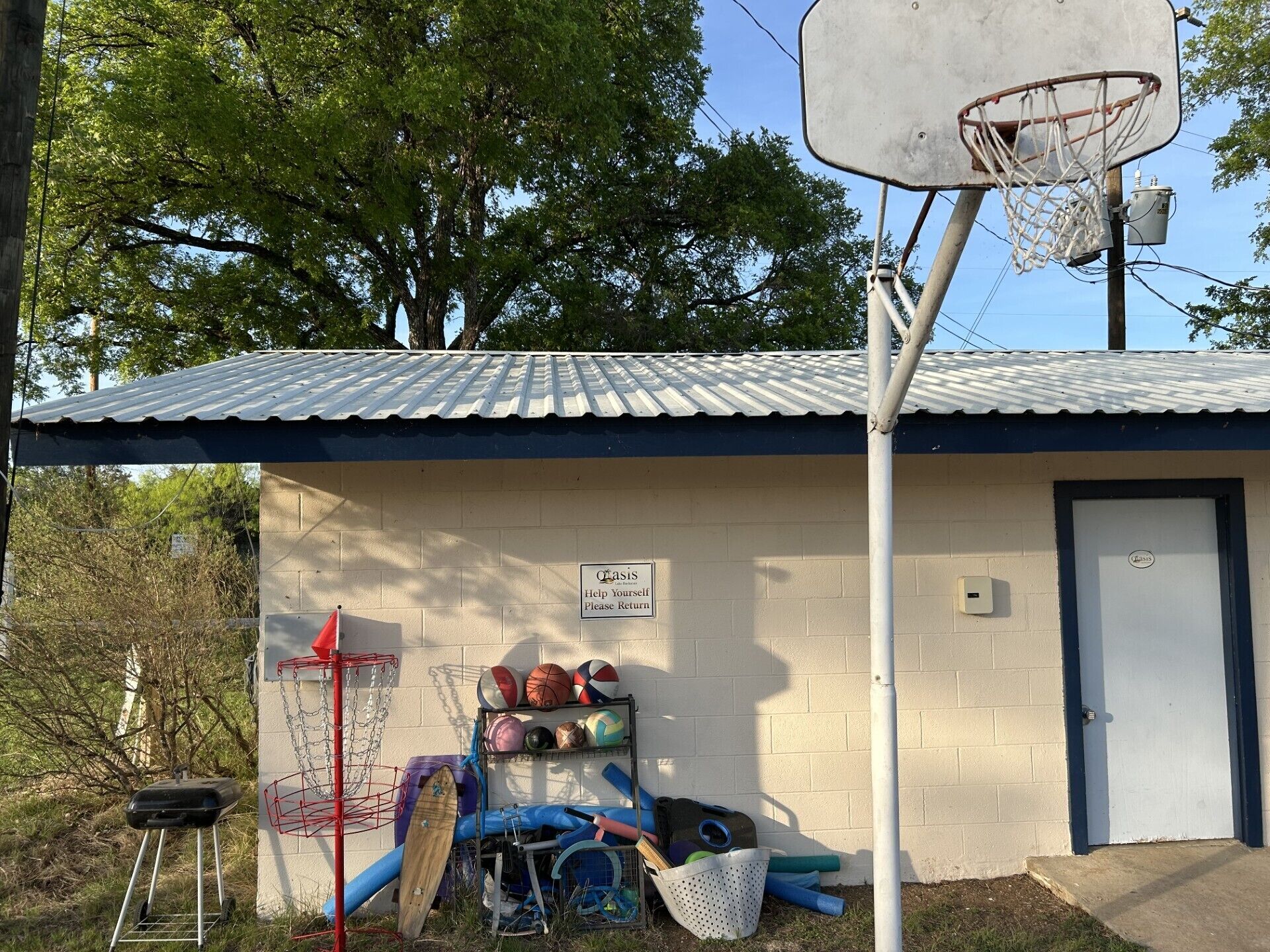 Un petit bâtiment avec un panier de basket, une pancarte indiquant « Merci de bien vouloir remettre les objets à leur place », et divers équipements sportifs, comme des ballons et une raquette.