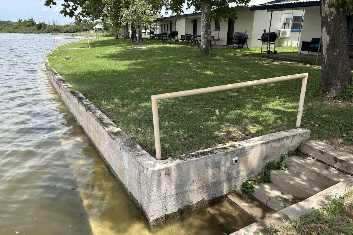 Een betonnen barrière met een metalen hekwerk langs water, een gazon met bomen en een gebouw met terras.