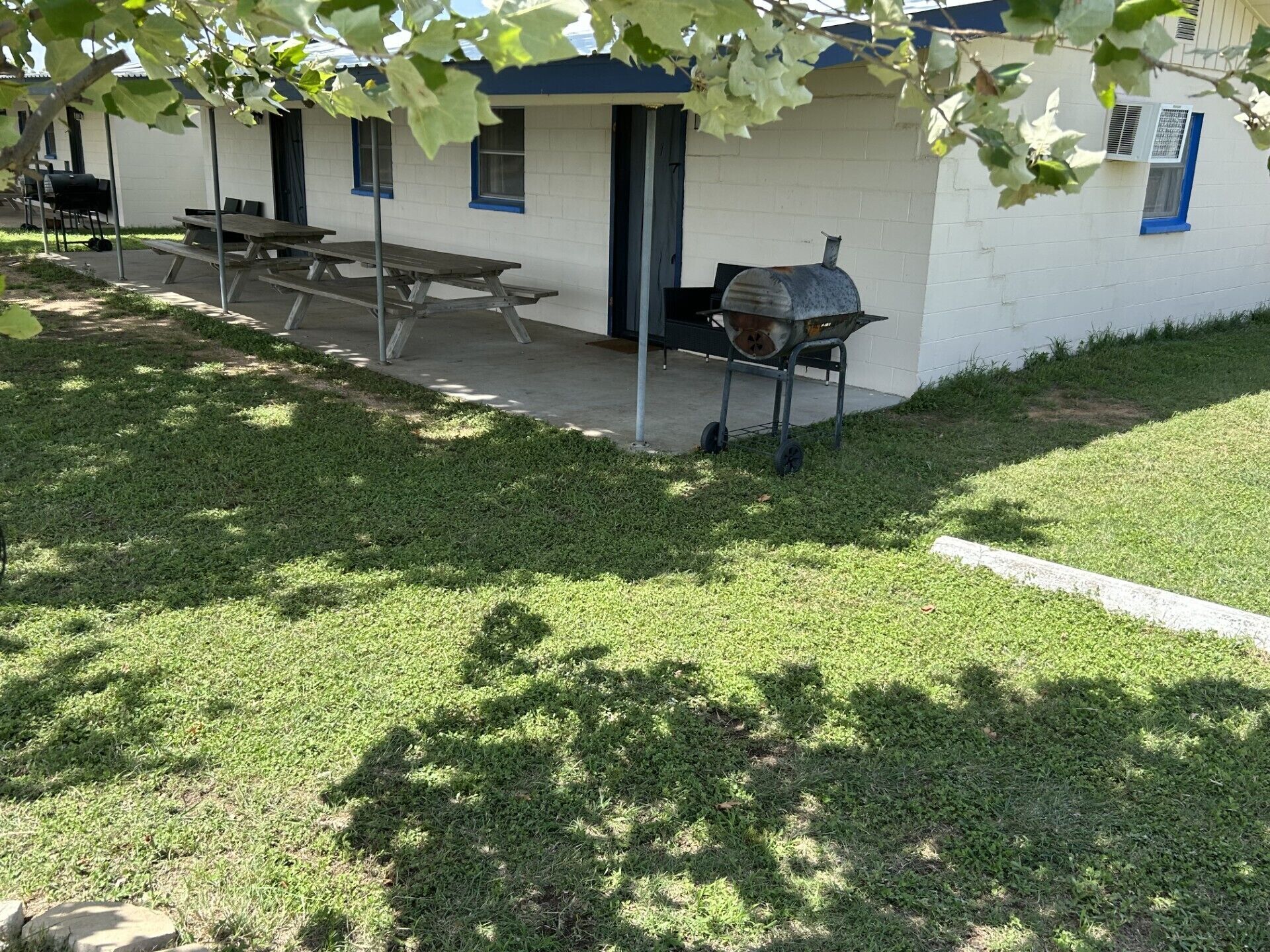 A patio area with picnic tables and a barbecue grill, surrounded by a well-maintained lawn and a building with a blue door.