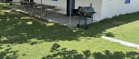 A patio area with picnic tables and a barbecue grill, surrounded by a well-maintained lawn and a building with a blue door.