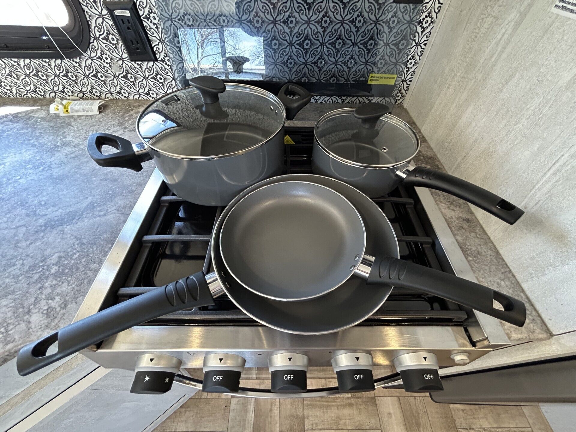 A kitchen stove with four pots and pans, a power outlet, and a window with a view of trees.