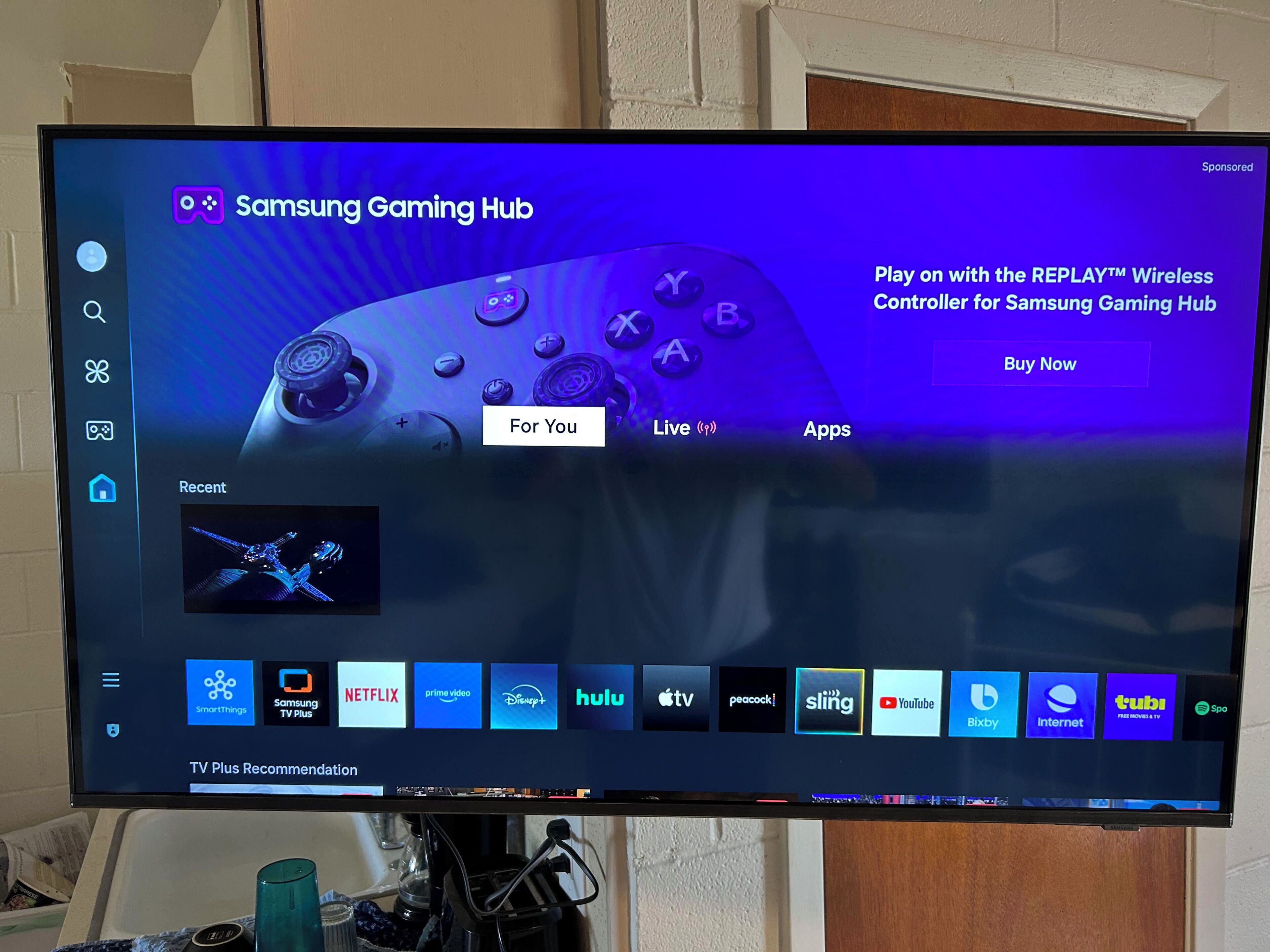 Ein Fernsehbildschirm zeigt die Samsung Gaming Hub-Oberfläche, mit einer Werbung für den REPLAY™ Wireless Controller.