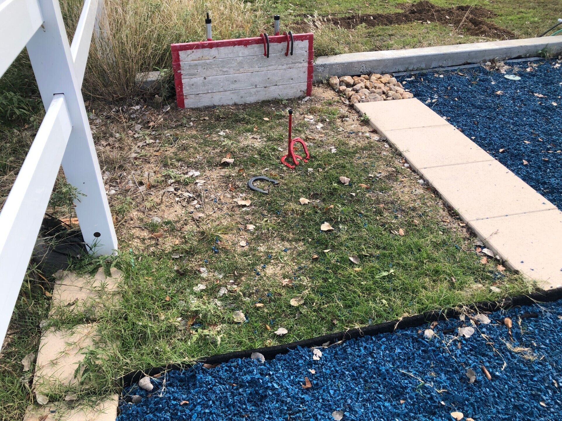 Een tuin met een witte metalen constructie, een rode en witte bank en een blauwe rubberen mat.