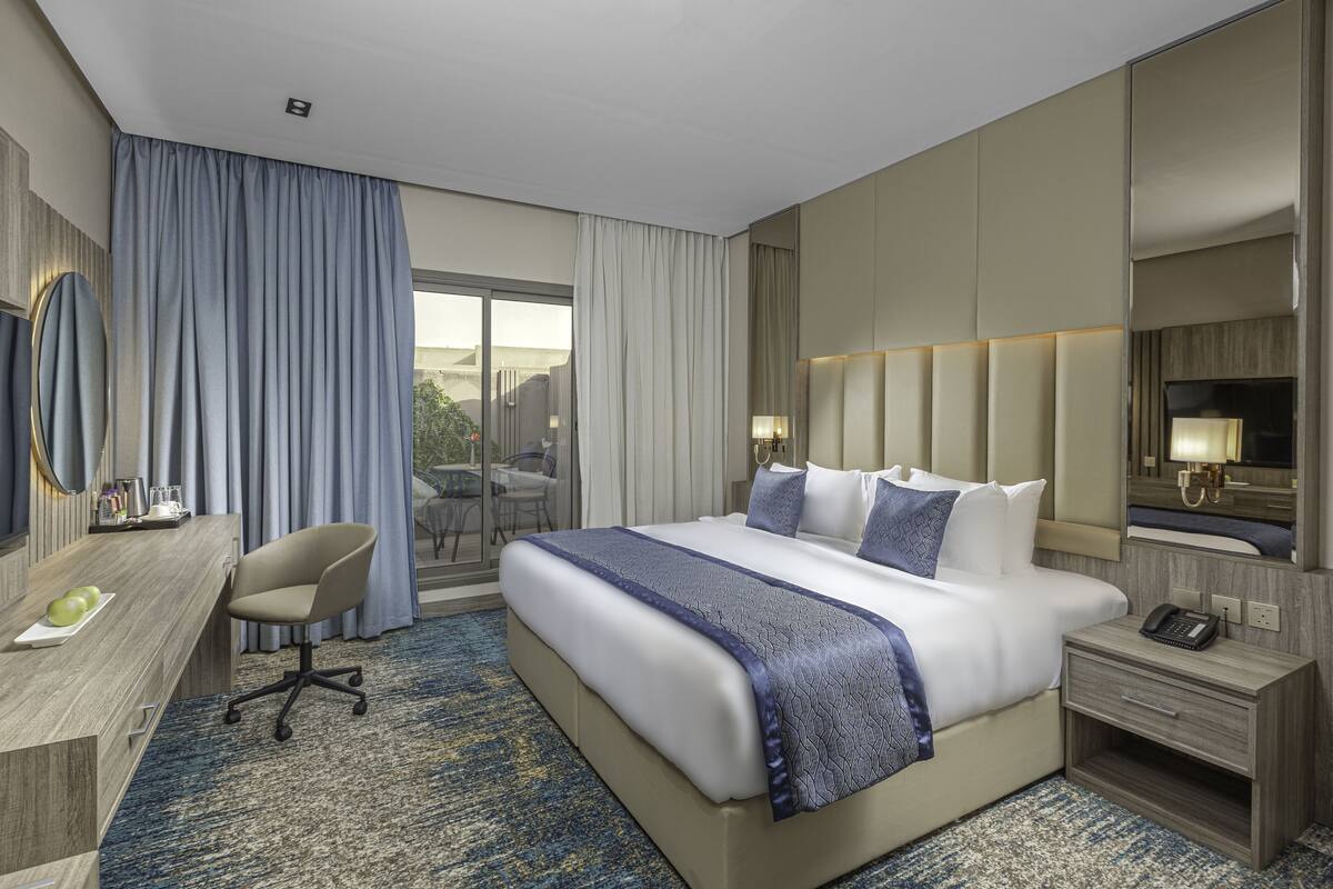 A hotel room with a large bed, a desk, a chair, a bedside table, a lamp, and a large window with curtains.