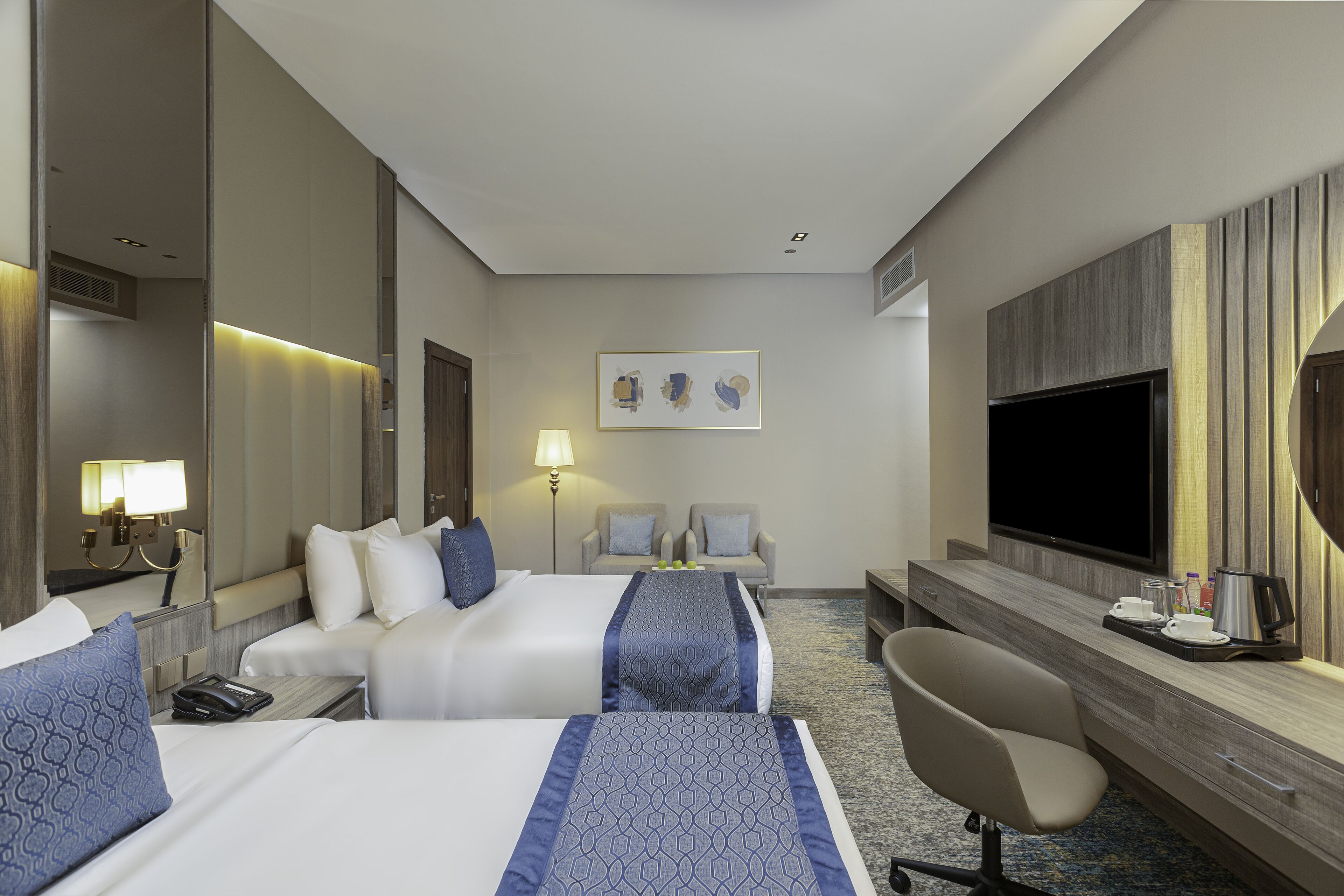A hotel room with two beds, a desk, a chair, a TV, and a painting on the wall.
