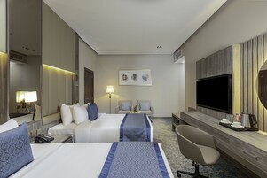 A hotel room with two beds, a desk, a chair, a TV, and a painting on the wall.