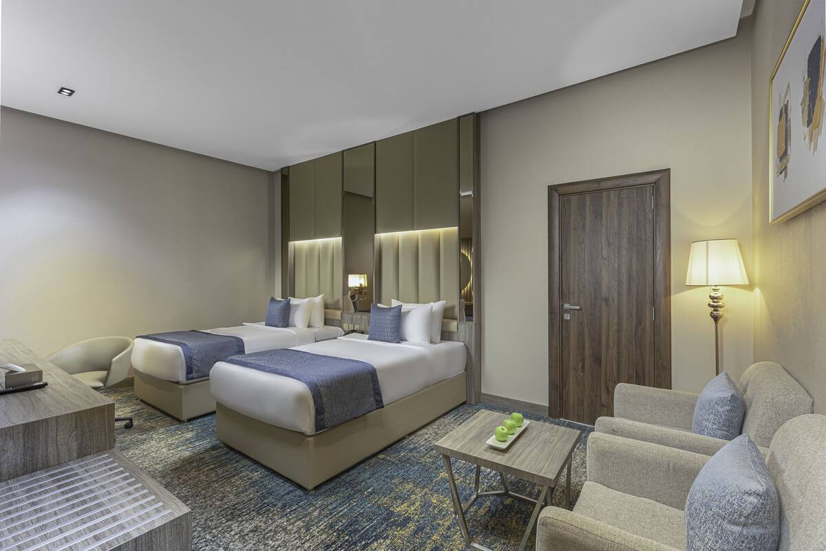 A hotel room with two beds, a desk, a chair, a sofa, a lamp, and a mirror.