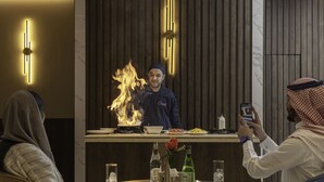 A restaurant setting with a chef performing a flambé, a dining table with plates, glasses, and a bottle of alcohol, and a person taking a photo. - Seven Gardens Hotel (Riyadh)
