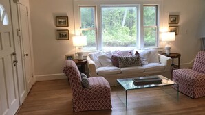 A living room with a white sofa, two patterned armchairs, a glass coffee table, and two side tables with lamps. There are three windows providing a view of greenery outside. - Renovated historic home! (Chapel Hill)