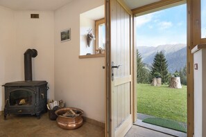 Una habitación con estufa de leña, una puerta que da a un espacio exterior cubierto de césped y montañas al fondo.