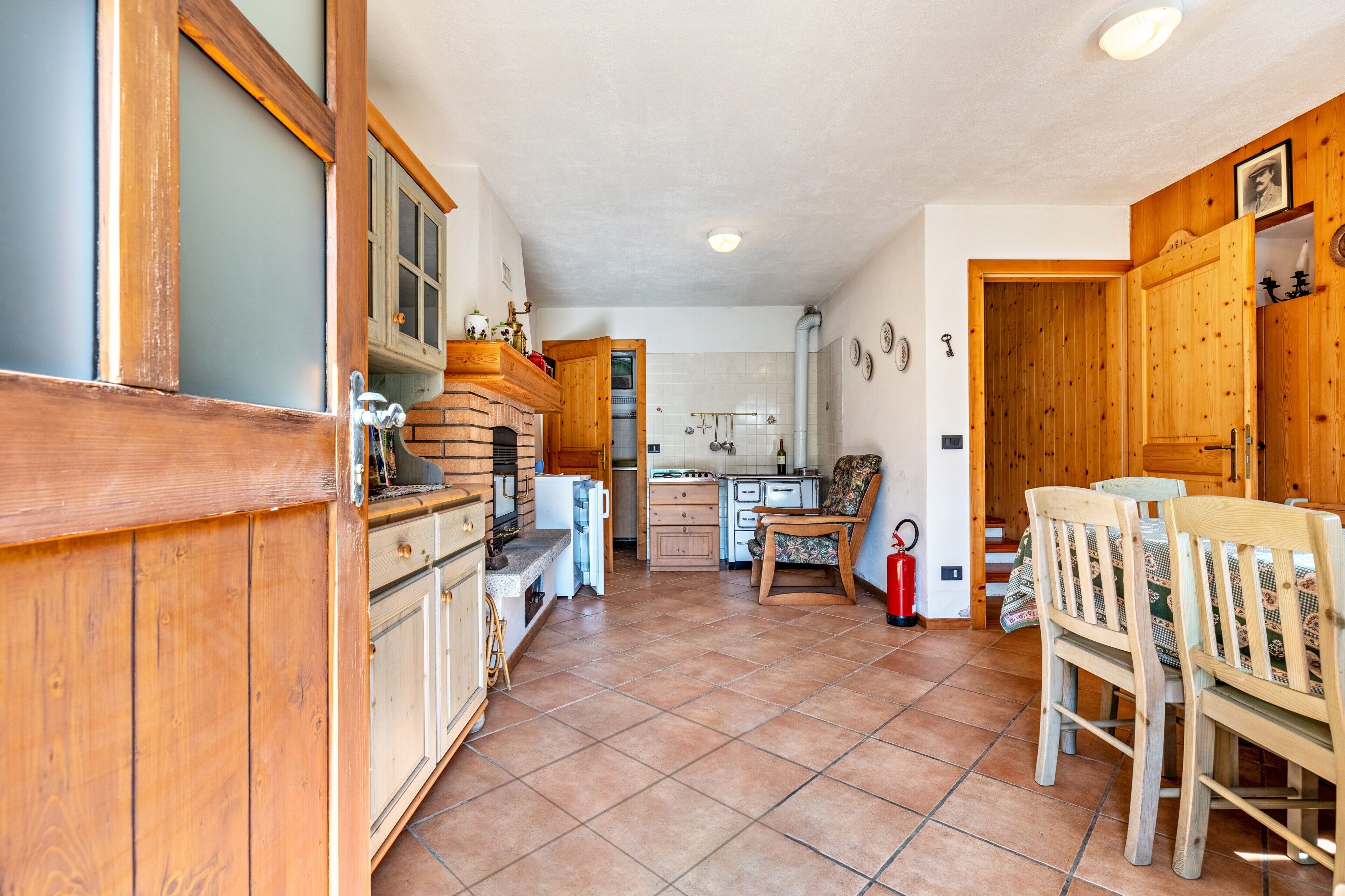 Une cuisine avec des meubles de rangement en bois, un sol carrelé, un coin repas avec des chaises et un réfrigérateur.