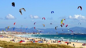 Una playa con muchos cometas, una costa y montañas a lo lejos.