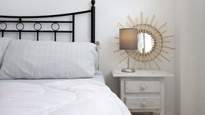 Una cama con edredón blanco y almohadas, cabecera de metal negro, mesita de noche blanca con cajones y un espejo con diseño de sol.