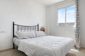 Una cama con colcha blanca, cabecero de metal negro y ventana con vista a un edificio.