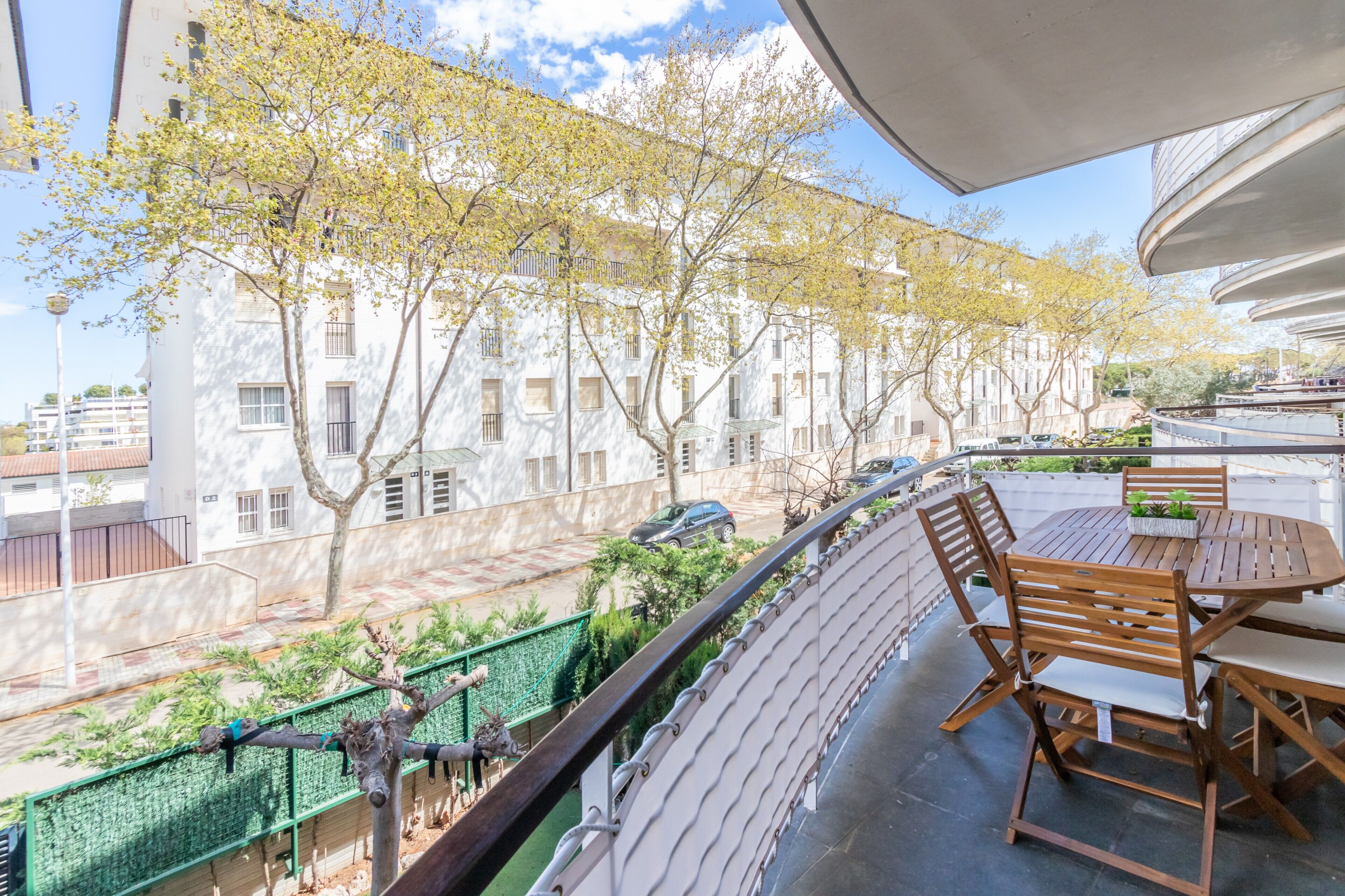 Un balcone con tavolo e sedie, affacciato su una strada con alberi e palazzi.