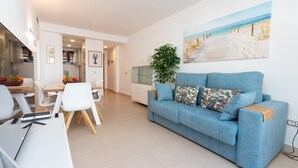 Un salon moderne avec un canapé bleu, un coin repas avec des chaises blanches, un espace cuisine et un tableau sur le thème de la plage accroché au mur.